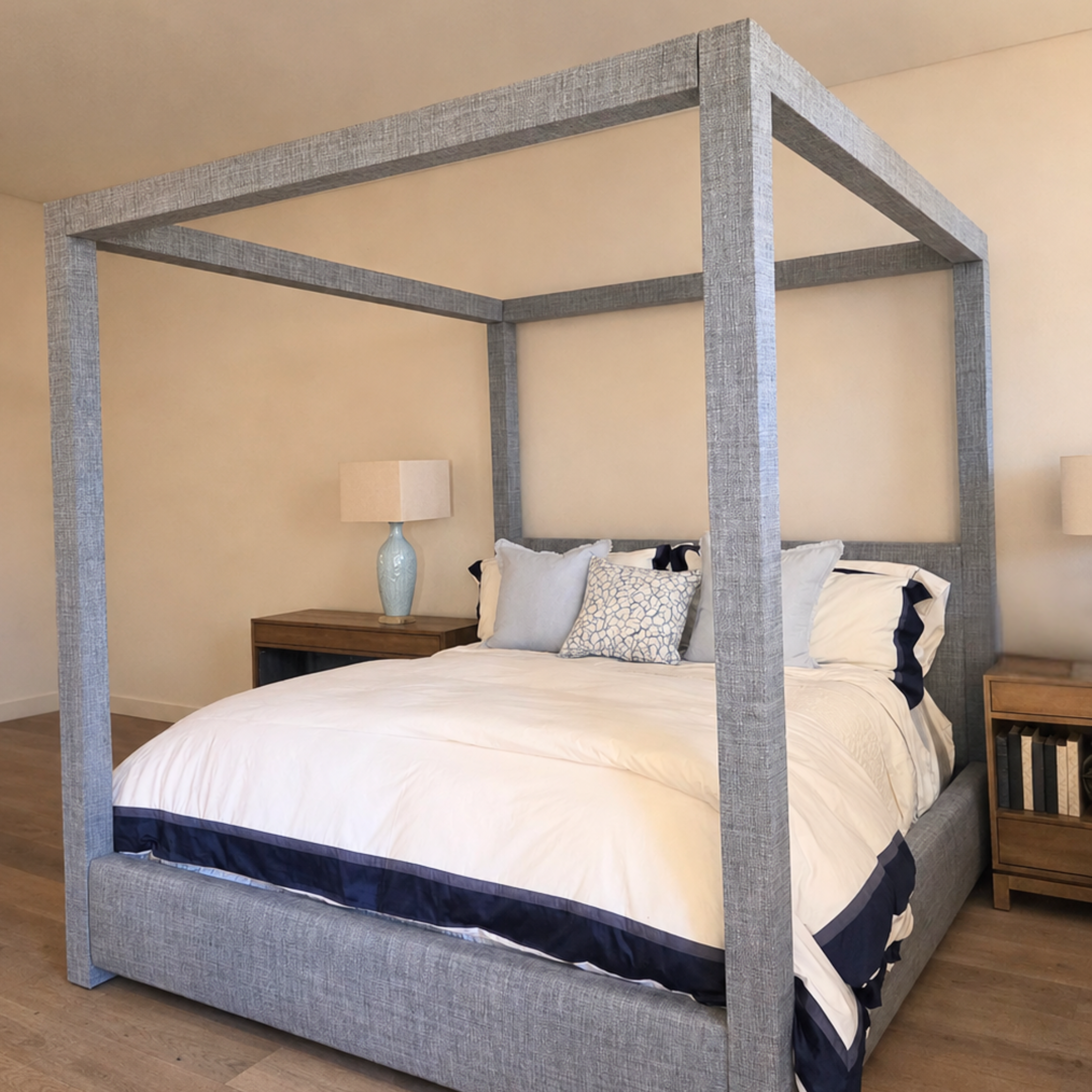 A four-poster canopy bed with a textured gray upholstered frame, dressed in white bedding with navy blue accents, set in a bedroom with wood flooring and bedside tables.