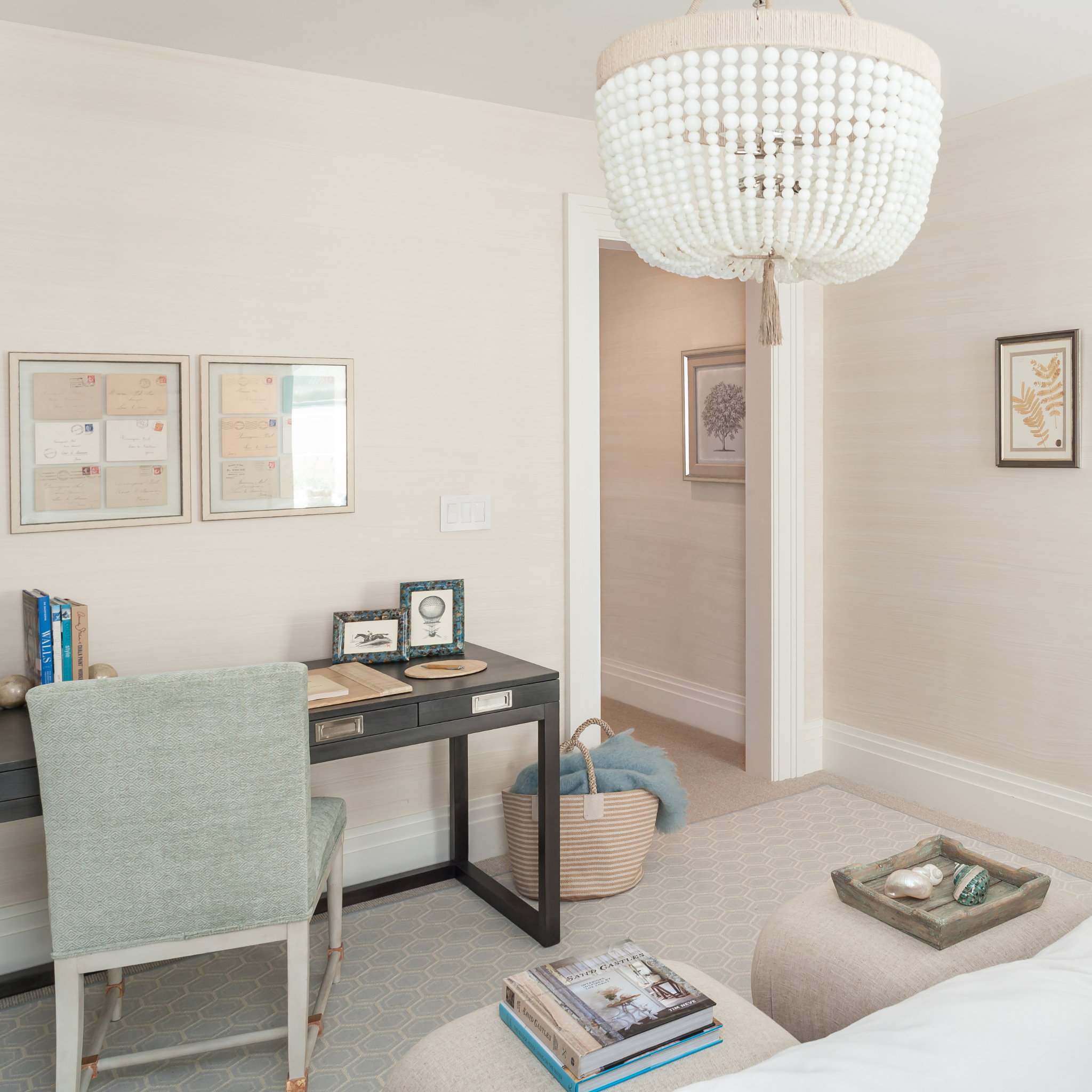 Neutral bedroom corner with a small desk, upholstered chair, and layered decor, accented by soft blues and a beaded chandelier overhead.