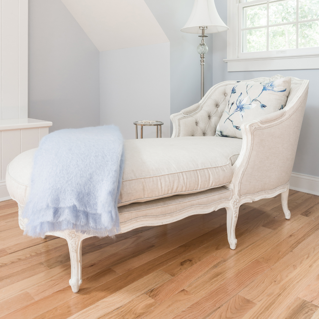 Beige upholstered bench with floral pillow and light blue throw blanket in a room with wooden floor and white walls.