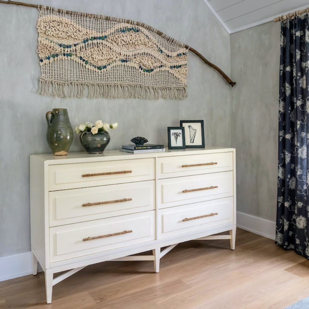 White dresser with gold handles in a room with a textured wall, decorative vase, and framed pictures.