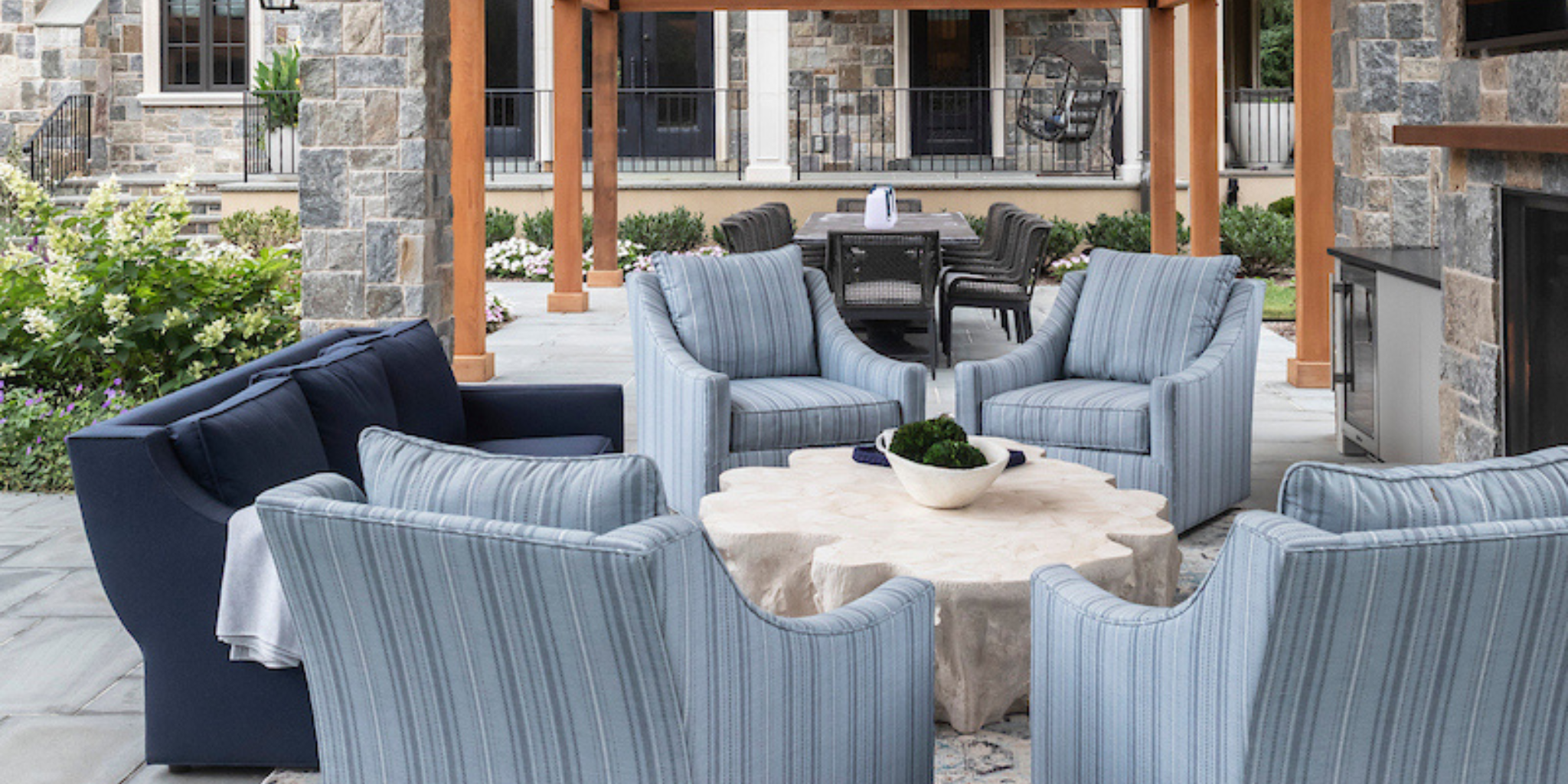 Outdoor patio with cushioned seating and a natural stone coffee table beneath a pergola, surrounded by lush greenery and a stone fireplace backdrop.