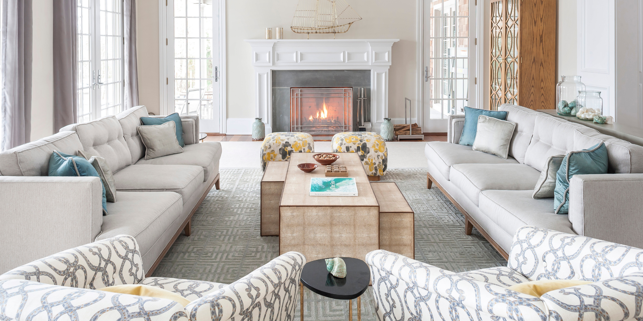 Light-filled living room with two sofas, patterned chairs, and a central coffee table, arranged around a white fireplace with soft blue and neutral accents.