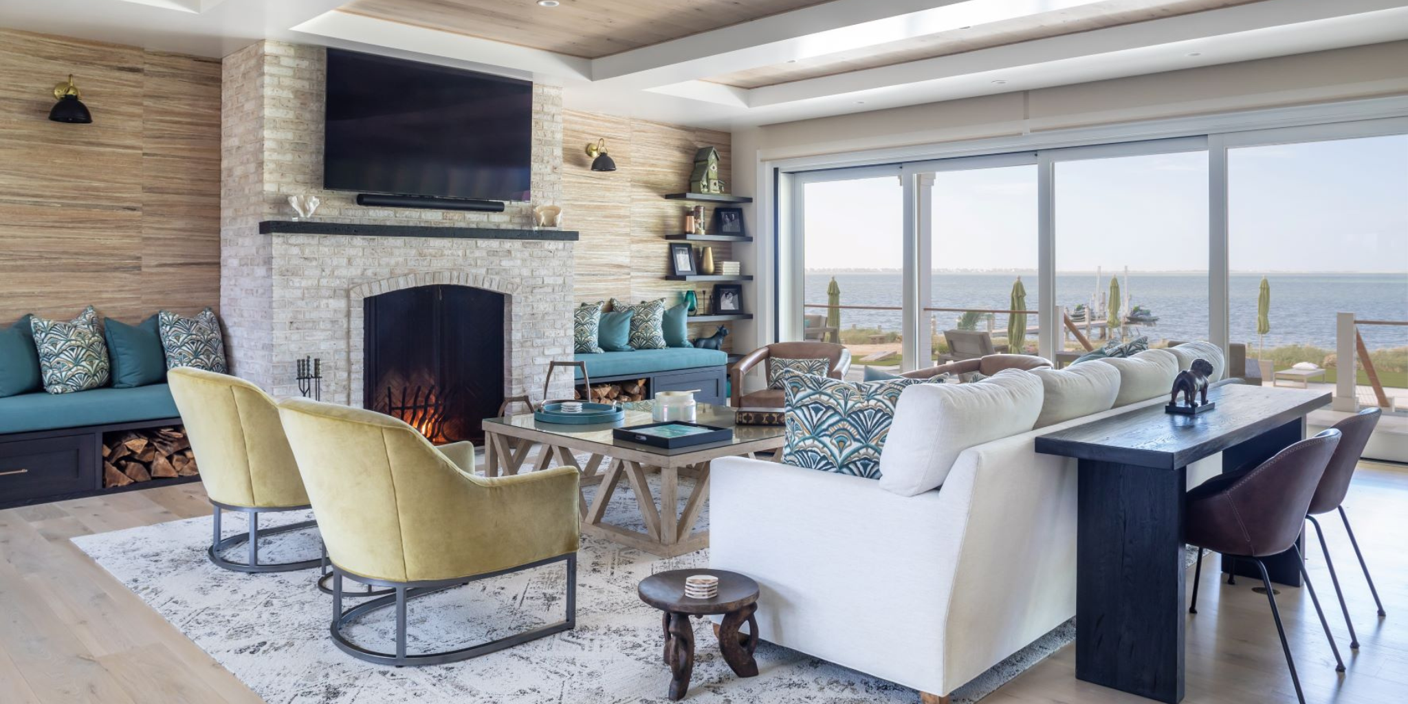 Open-concept coastal living room with a stone fireplace, layered seating, and large sliding doors opening to ocean views, styled with soft blues and natural textures.