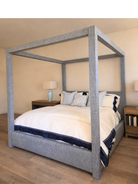 A four-poster canopy bed with a textured gray upholstered frame, dressed in white bedding with navy blue accents, set in a bedroom with wood flooring and bedside tables.