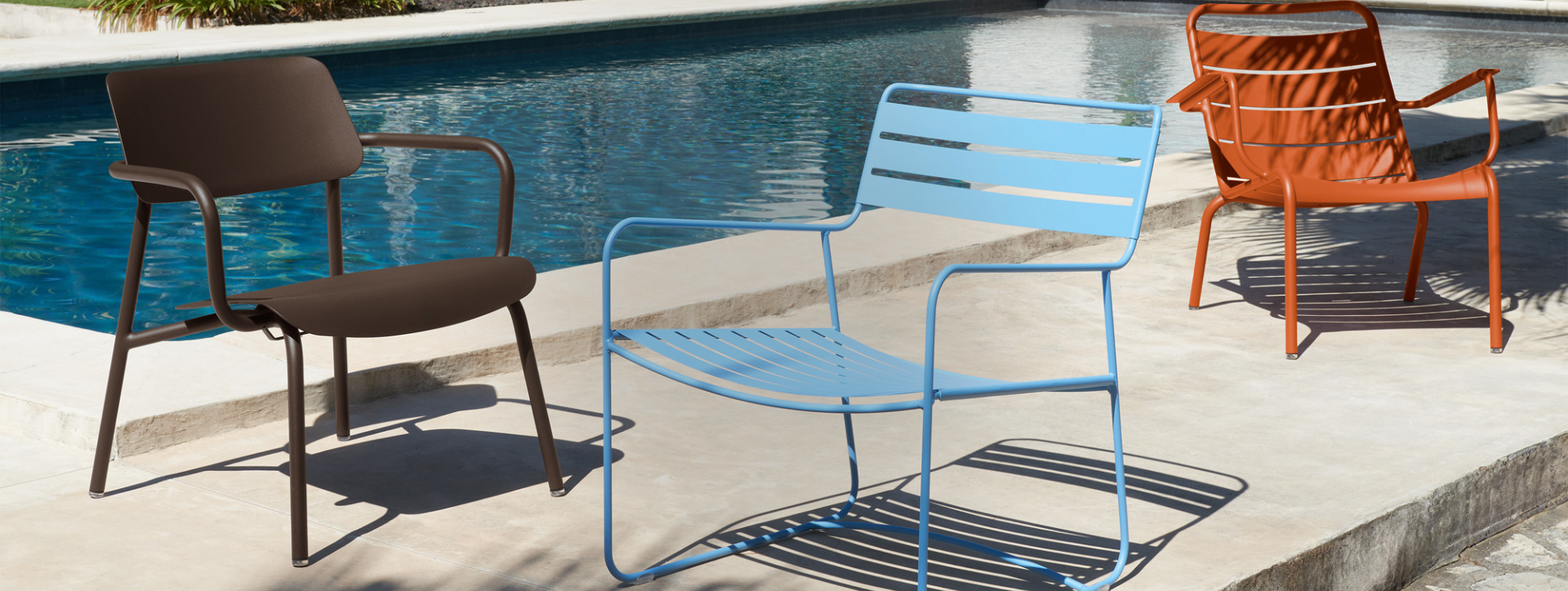Three outdoor chairs in brown, blue, and orange by a pool.