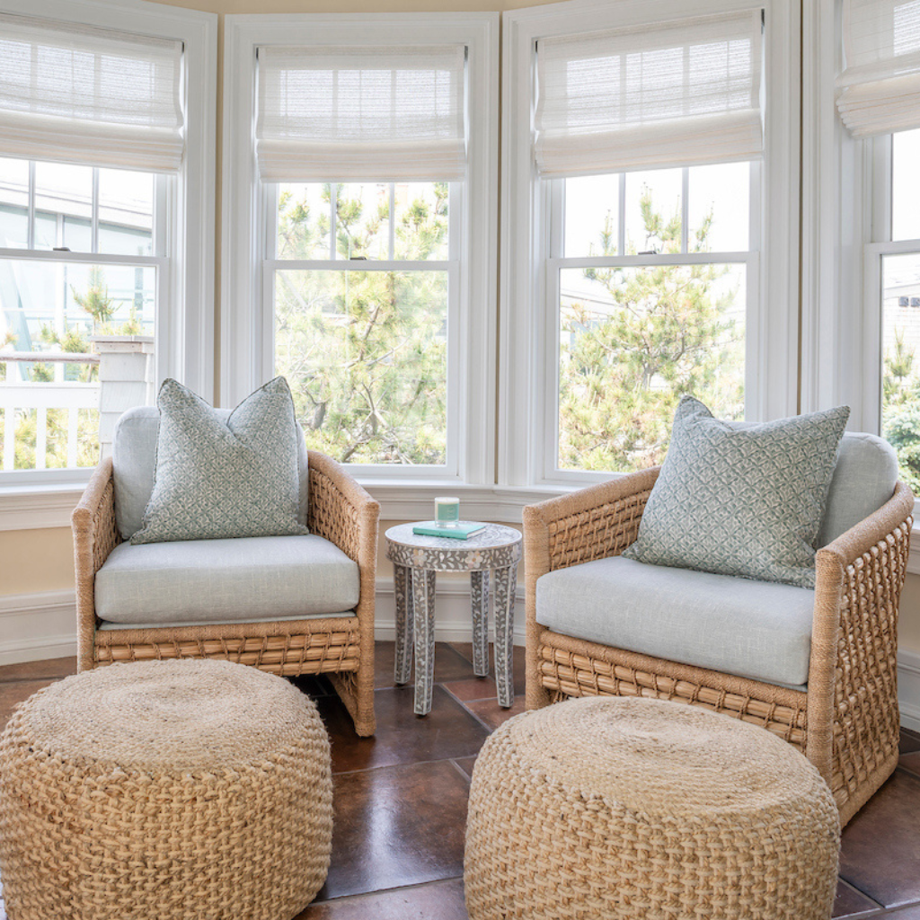 Wicker chairs with cushions and ottomans in a sunroom with large windows.