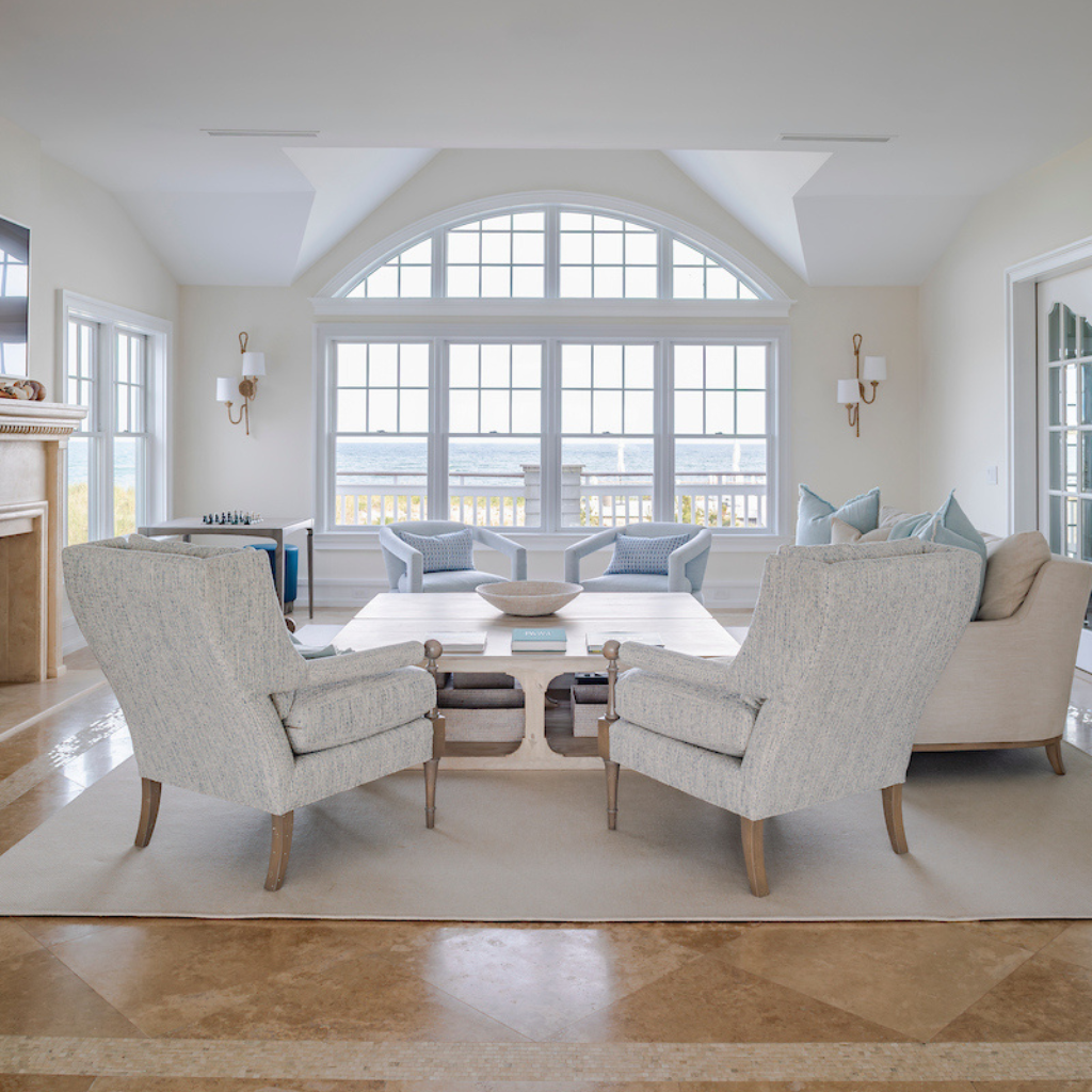 Living room with beige chairs, coffee table, and couches in a bright room with large windows.