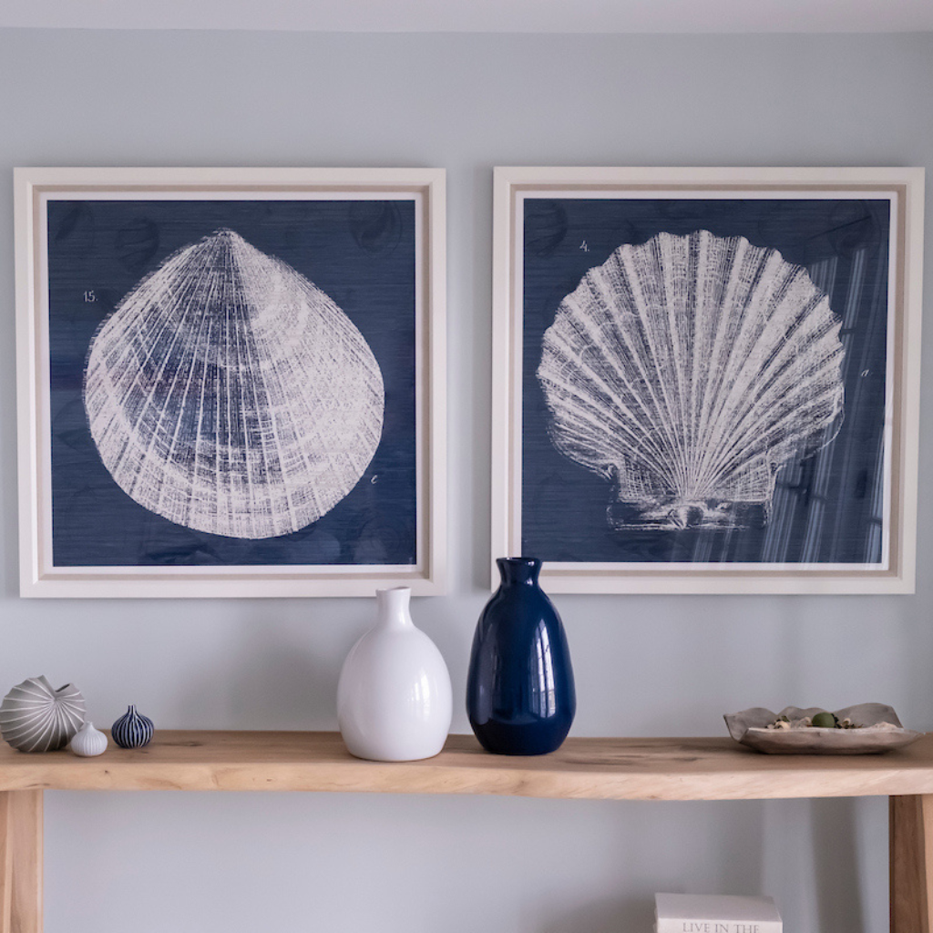 Two framed shell prints on a wall above a wooden shelf with decorative vases and items.