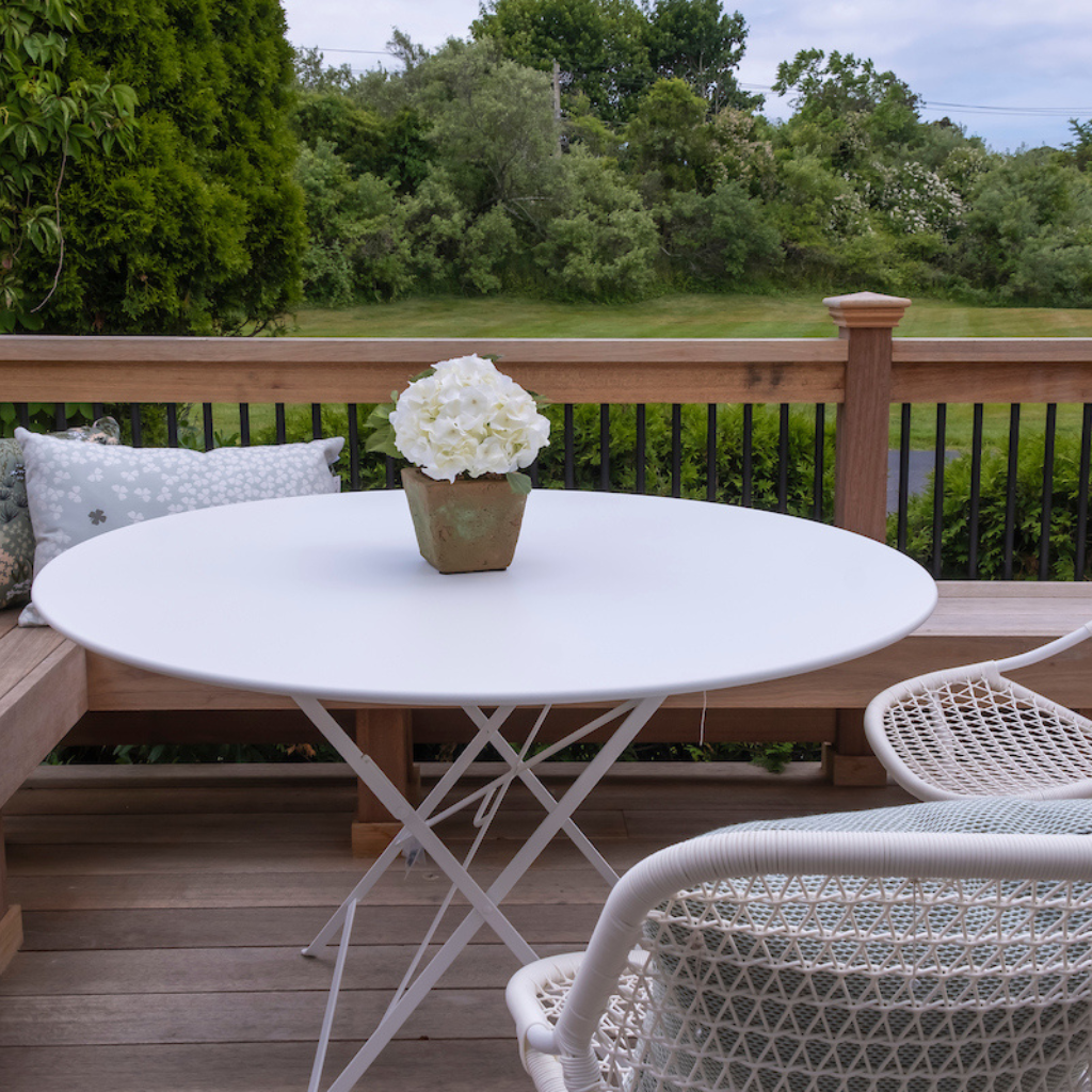White round table with a vase of flowers on a wooden deck with chairs and a view of trees.