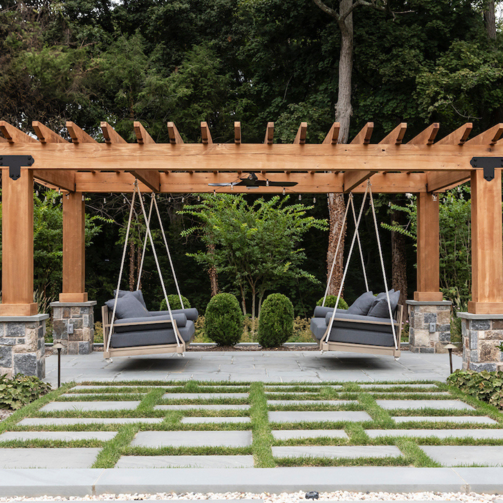 Two swing chairs under a wooden pergola in a garden setting with greenery.