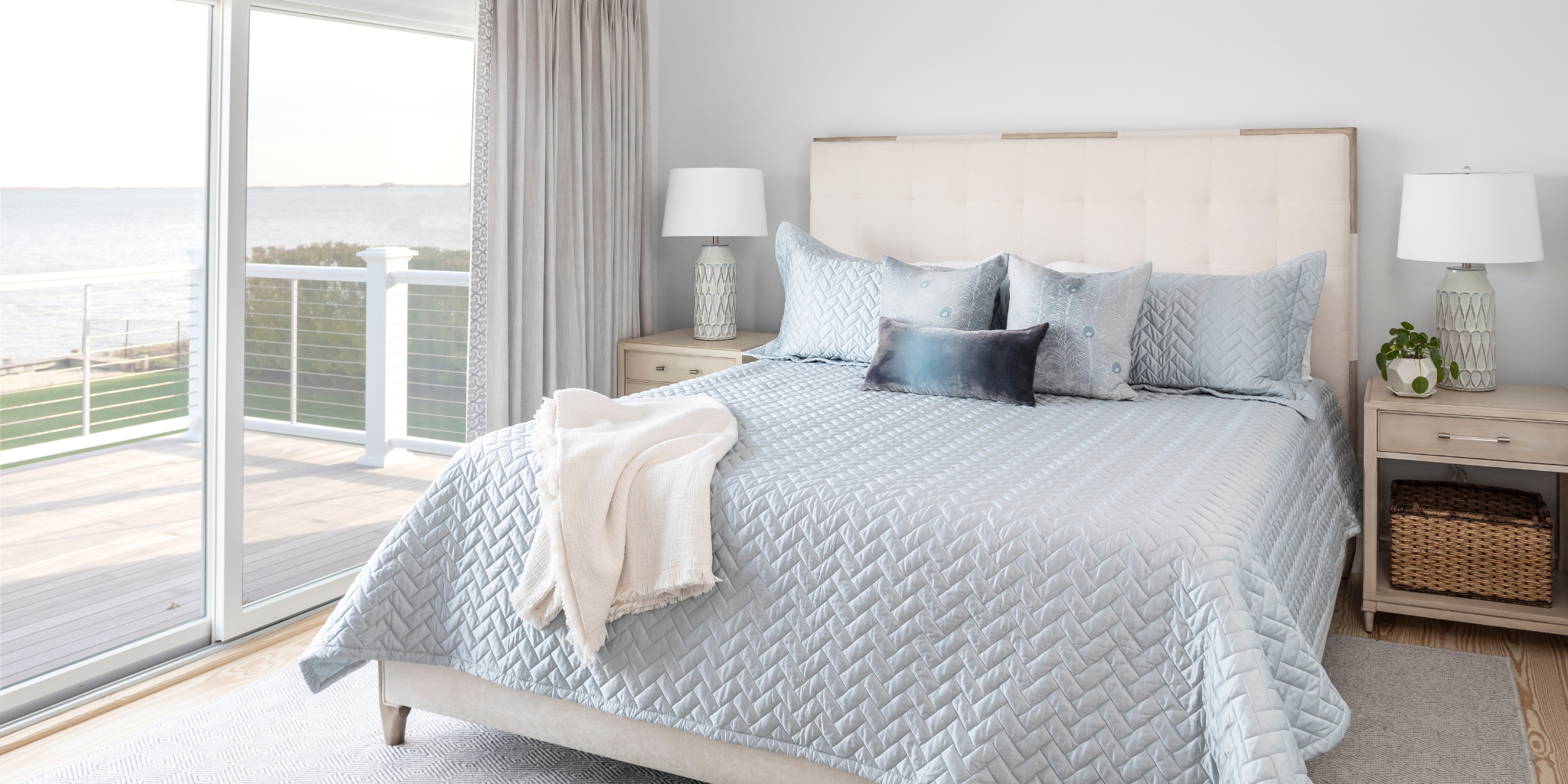 Bedroom with a bed covered in light blue bedding, nightstands with lamps, and a view of a garden.