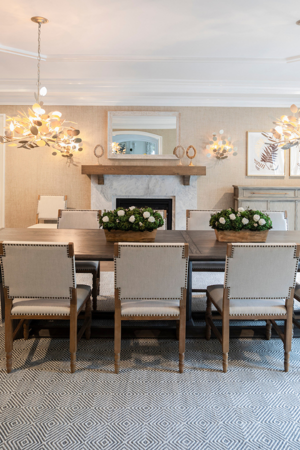 Refined dining room with a long wood table, upholstered nailhead chairs, and a fireplace backdrop, accented by sculptural lighting and greenery centerpieces.
