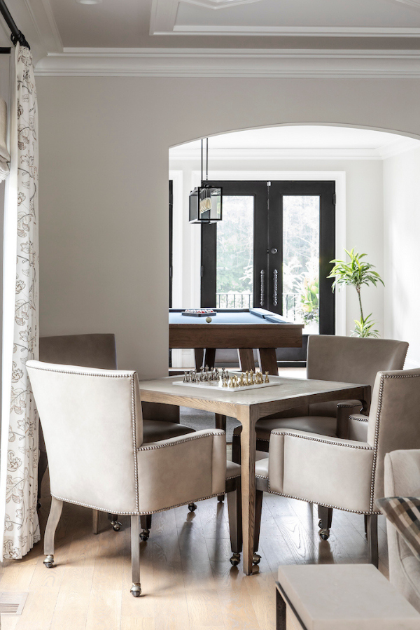 Neutral game area with a wood table and upholstered chairs, styled with a chess set, opening to a view of a pool table and glass doors beyond.