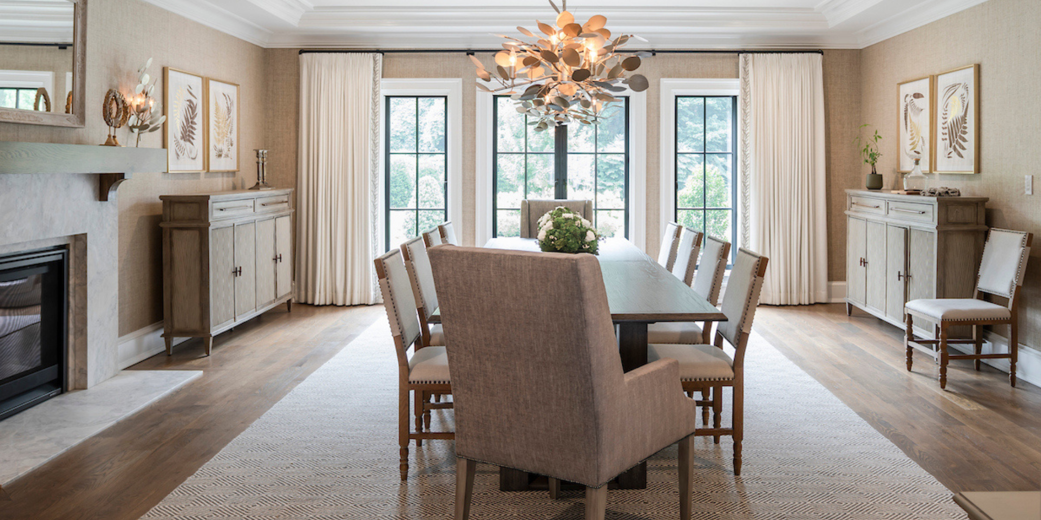 Neutral dining room with a long wooden table, upholstered chairs, and a sculptural chandelier, framed by large windows, linen drapery, and matching sideboards.