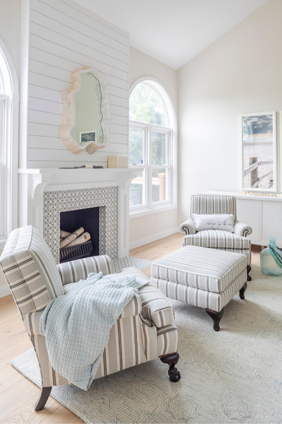 Light-filled sitting area with striped armchairs and ottoman, a white fireplace with decorative tile, and soft coastal accents near an arched window.