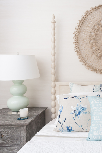 Neatly made bed with decorative pillows, nightstand with lamp and candle, and decorative wall hanging.