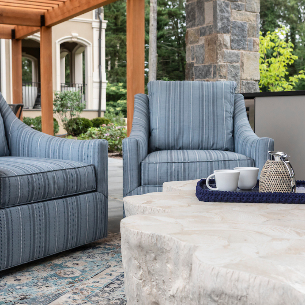Outdoor patio seating with blue upholstered chairs and a natural wood coffee table, styled with a tray, cups, and a woven lantern beneath a covered pergola.