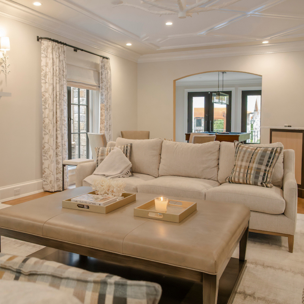 Cozy living room with a neutral sofa and oversized ottoman, opening to a game room beyond.