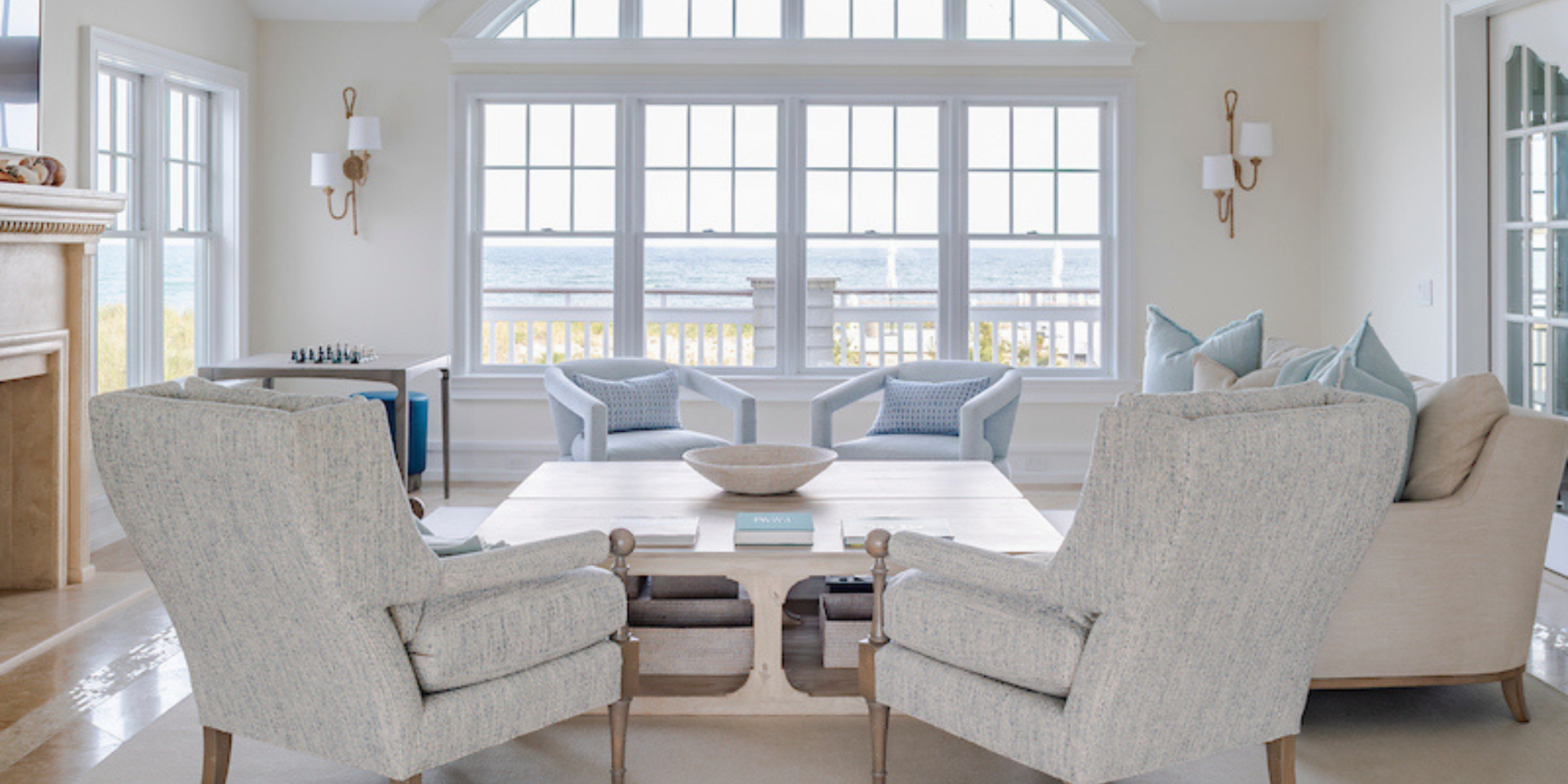 Bright coastal living room with neutral seating, a central coffee table, and large windows overlooking the ocean, styled with soft blue accents and natural textures.