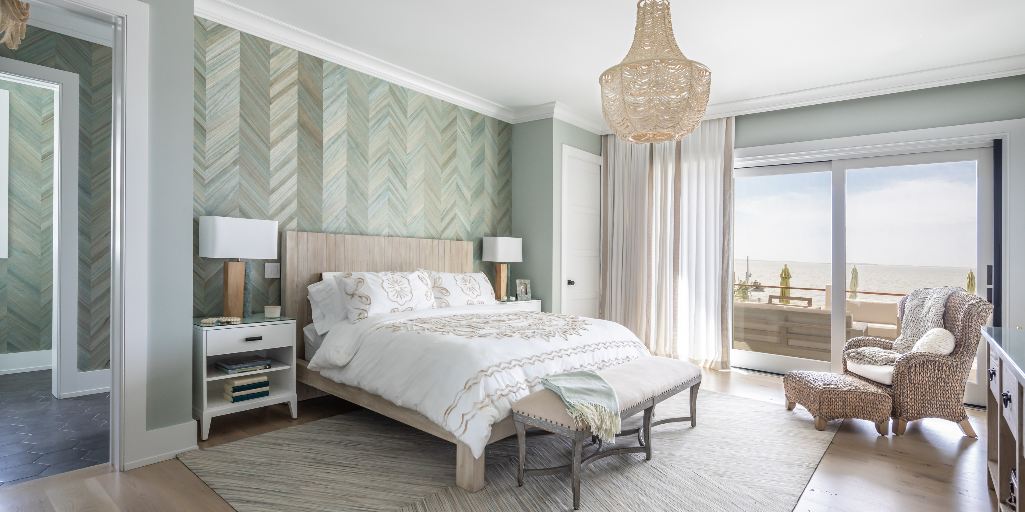 Coastal bedroom with a wood bed, soft neutral bedding, chevron-patterned wallpaper, and a woven chair, opening to a balcony with ocean views.