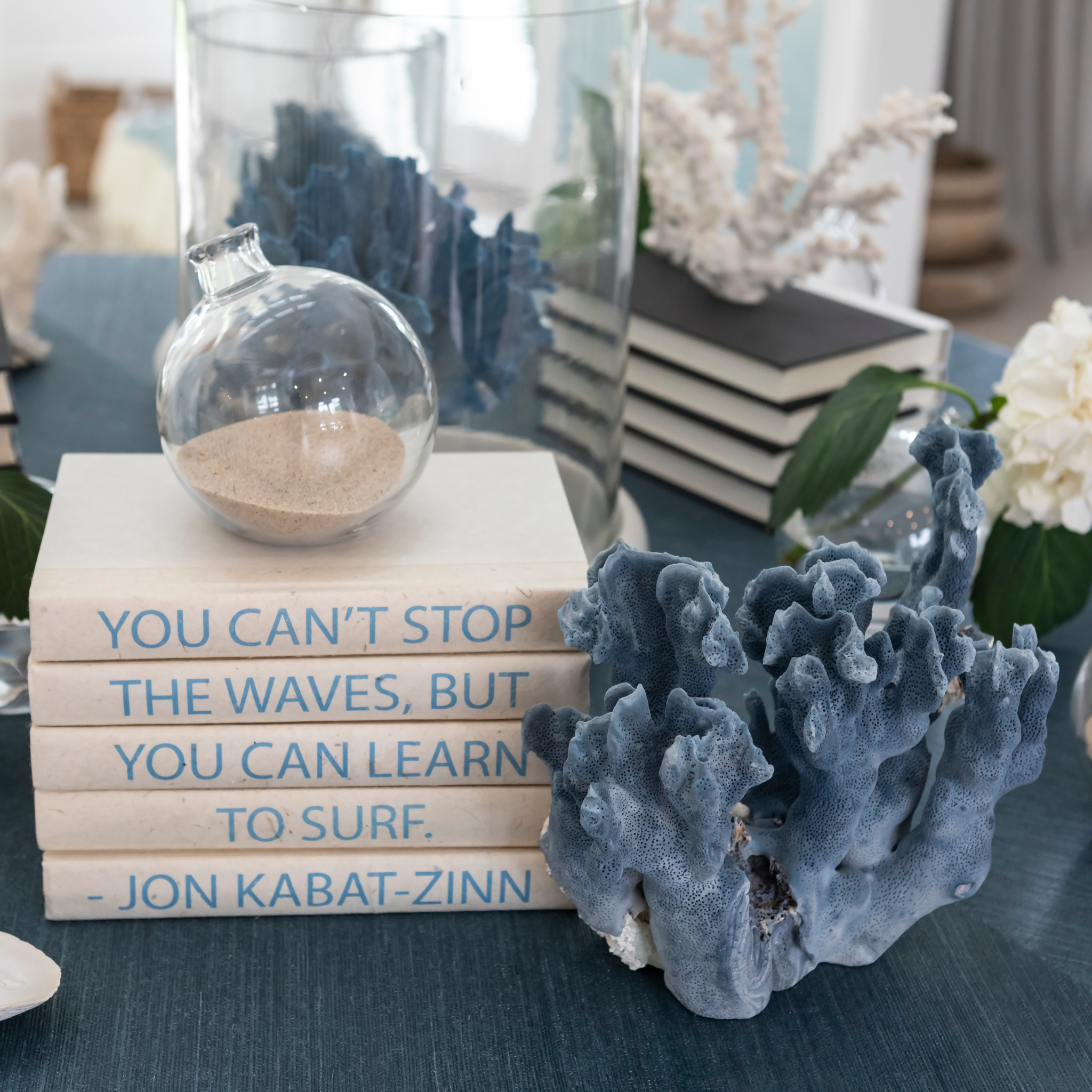 Coastal tabletop styling with coral decor, stacked books featuring a surf quote, and glass vessels arranged in soft blue and neutral tones.