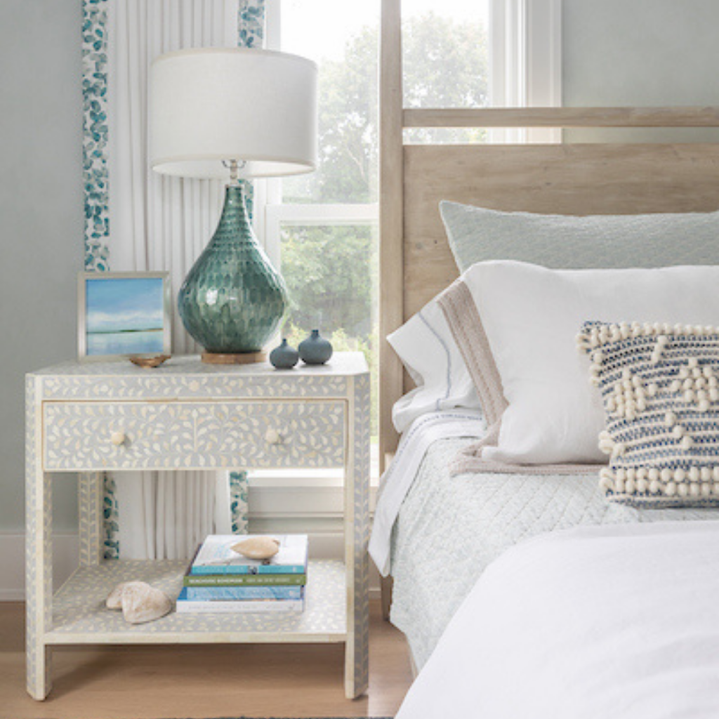 Light, coastal-inspired bedroom vignette with a carved nightstand, ceramic table lamp, layered bedding, and soft blue accents by a sunlit window.